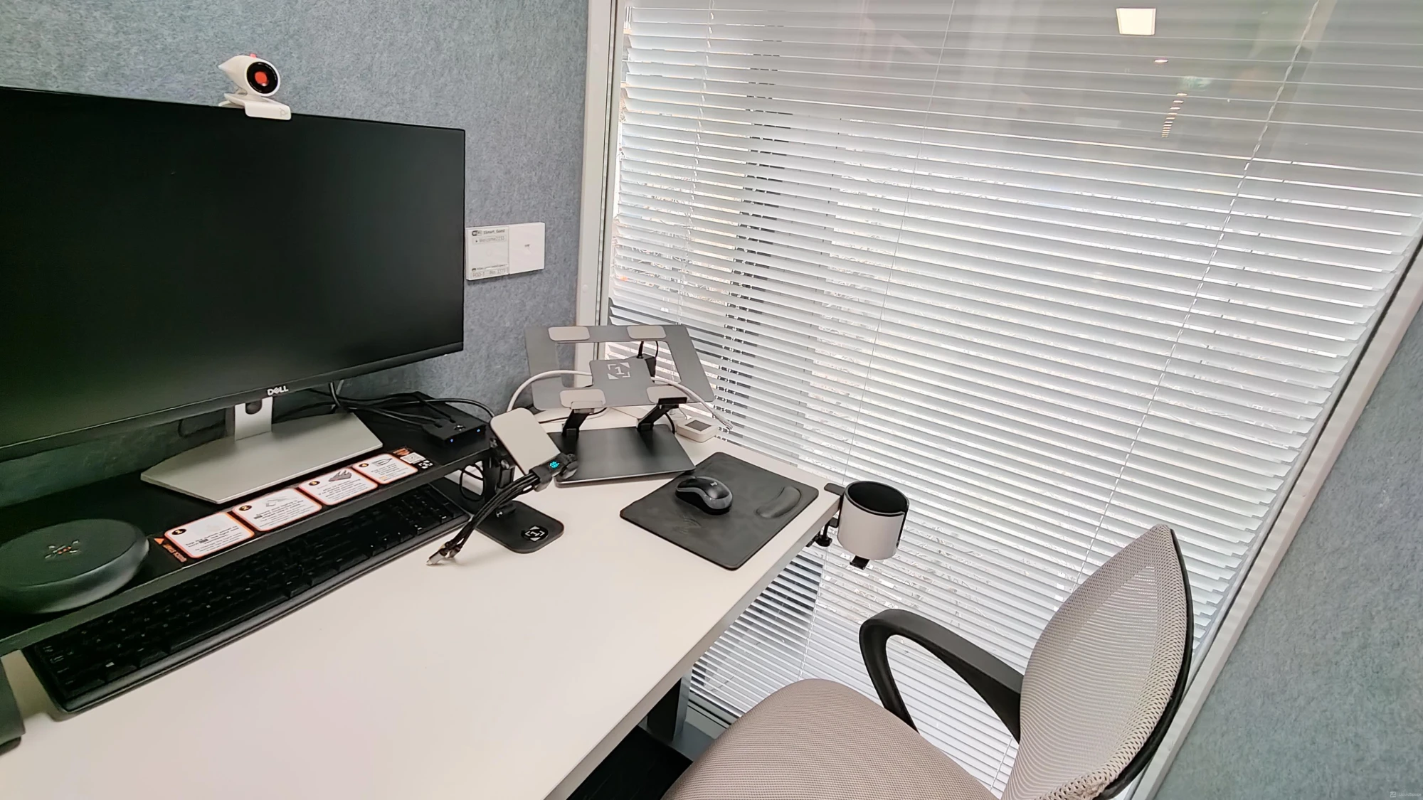 A SmartVue interview desk: monitor with webcam, keyboard, mouse and laptop stand beside a window with blinds
